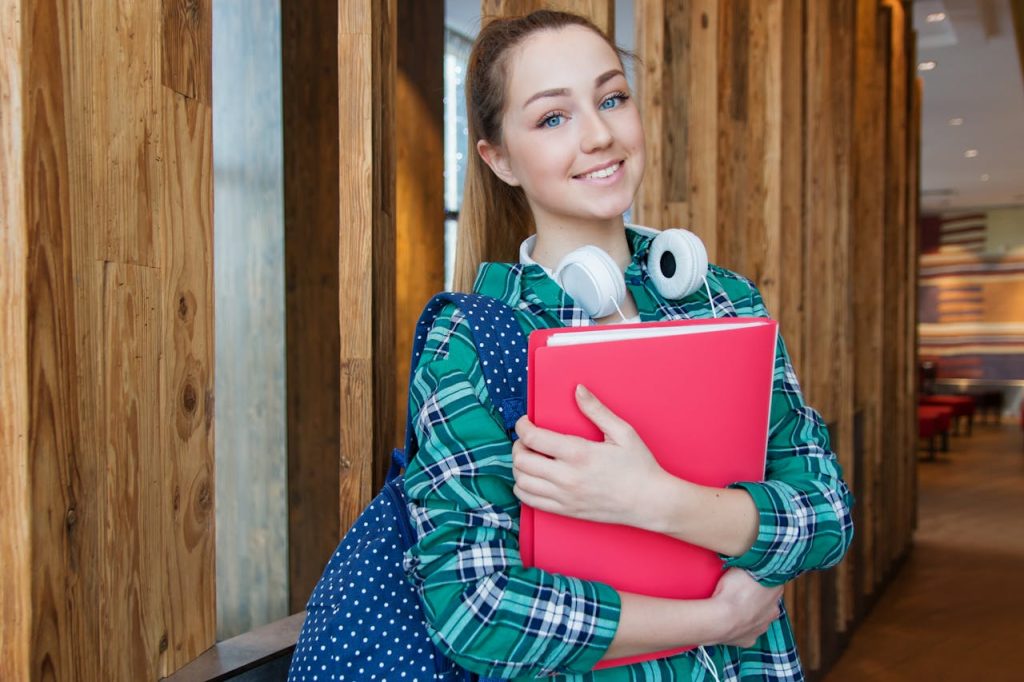 woman-standing-in-hallway-while-holding-book-1462630 Smiling student holding book and wearing headphones, ready for study.